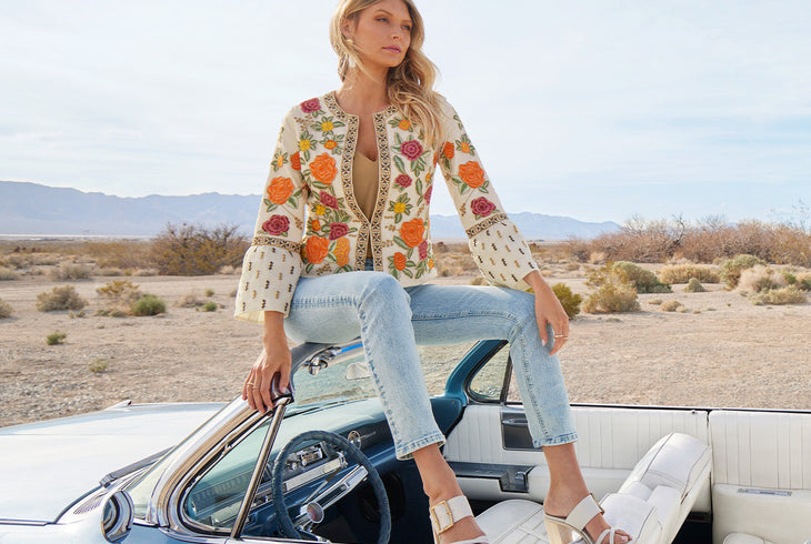 Model wearing a floral embroidered flare-sleeve jacket, tan v-neck charmeuse blouse, light wash jeans, and white buckle heels while sitting on a vintage convertible.